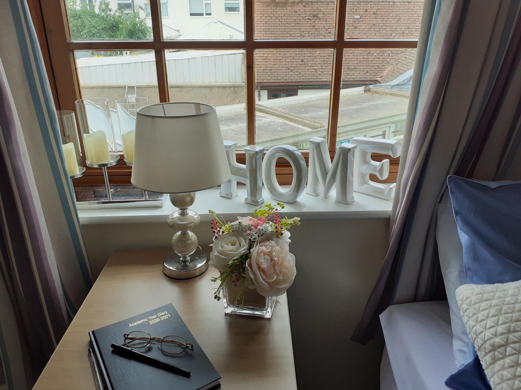 side table holding flowers, lamp and a diary next to a window